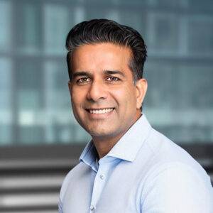 Portrait of Lalit Ahluwalia, smiling, wearing a light blue shirt in front of a blurred office window
